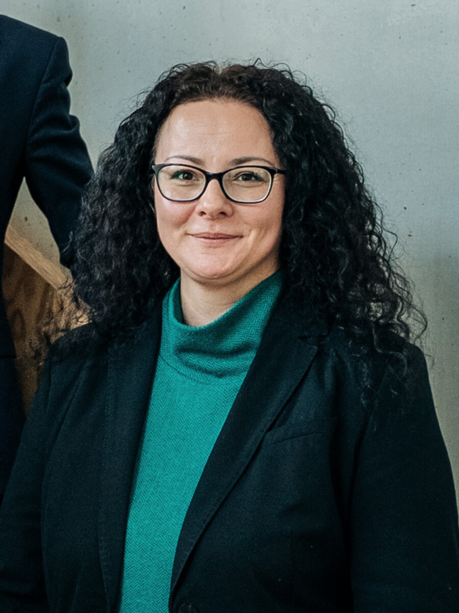 Eine Frau mit lockigem dunklem Haar und Brille, die einen blaugrünen Rollkragenpullover und einen schwarzen Blazer trägt, lächelt in die Kamera vor einem neutralen Hintergrund.