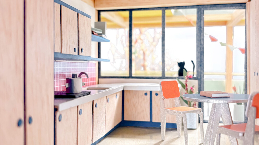 Miniaturküche mit hellen Holzschränken, rosa Fliesenspiegel und rundem Tisch. Eine schwarze Katze sitzt auf der Fensterbank und schaut nach draußen. Sonnenlicht strömt durch die großen Fenster, an denen festliche Wimpelketten hängen.