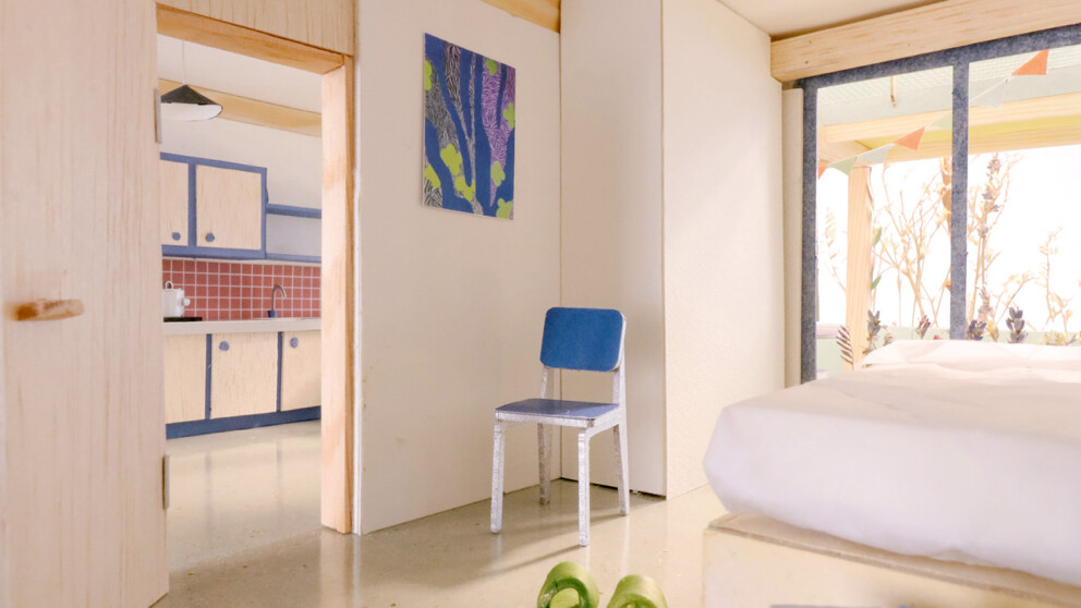 Ein aufgeräumtes Schlafzimmer mit einem blauen Stuhl, grünen Hausschuhen, einem Bett und Blick in die Küche. Helles Sonnenlicht strömt durch große Glasschiebetüren herein.