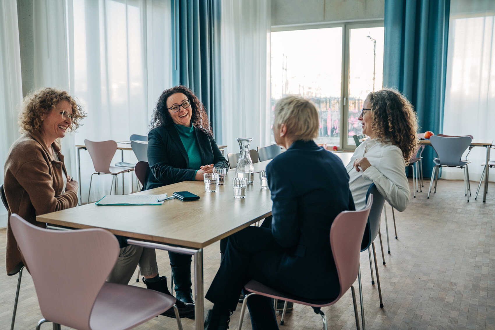 Vier Frauen sitzen um einen Tisch in einem hellen, modernen Raum, lächeln und unterhalten sich, vor ihnen stehen Gläser mit Wasser und Papiere.