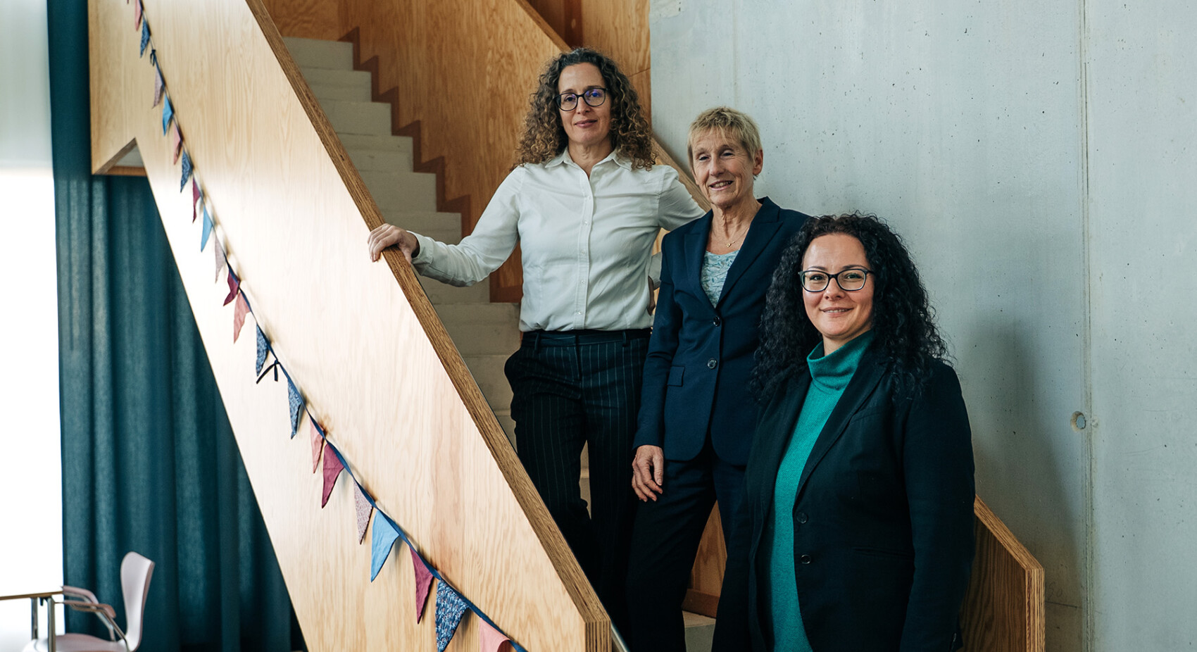 Drei Frauen in Geschäftskleidung posieren gemeinsam auf einer mit bunten Wimpeln geschmückten Holztreppe in einem modernen Innenbereich mit Betonwänden.
