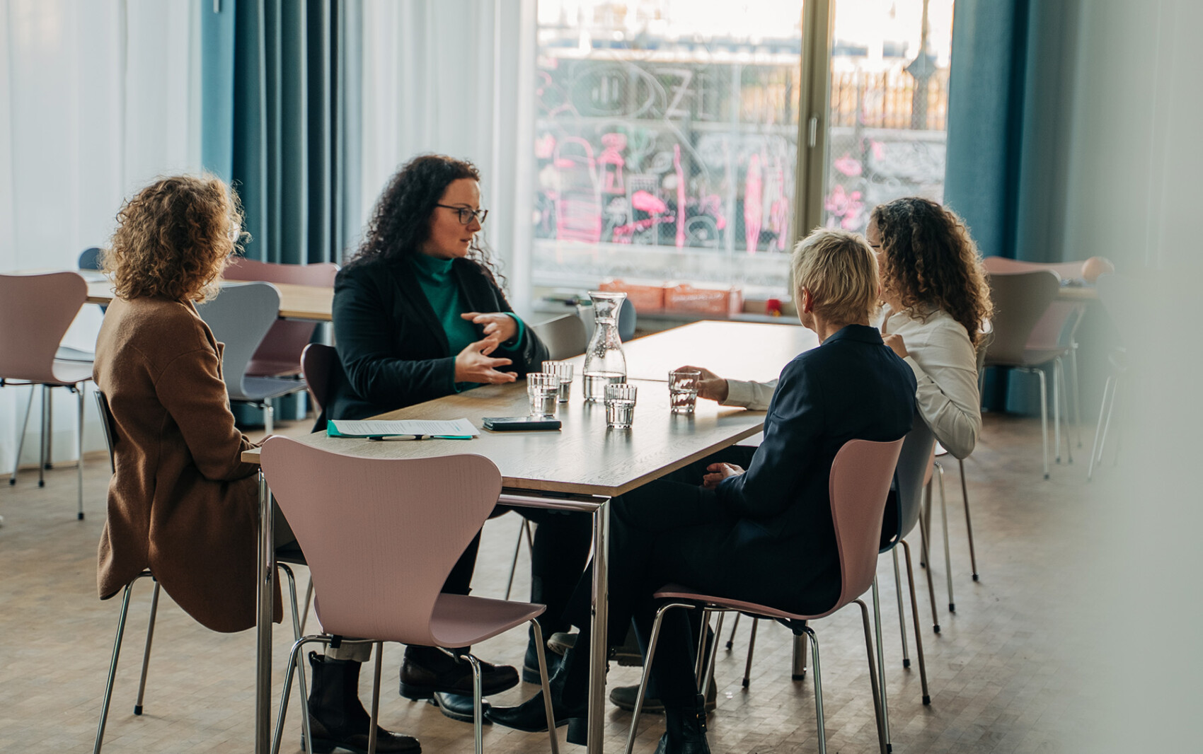 Vier Personen sitzen in einem modernen Büro um einen Tisch herum und führen eine Diskussion. Auf dem Tisch stehen Wassergläser und ein Krug. Natürliches Licht fällt durch große Fenster im Hintergrund.