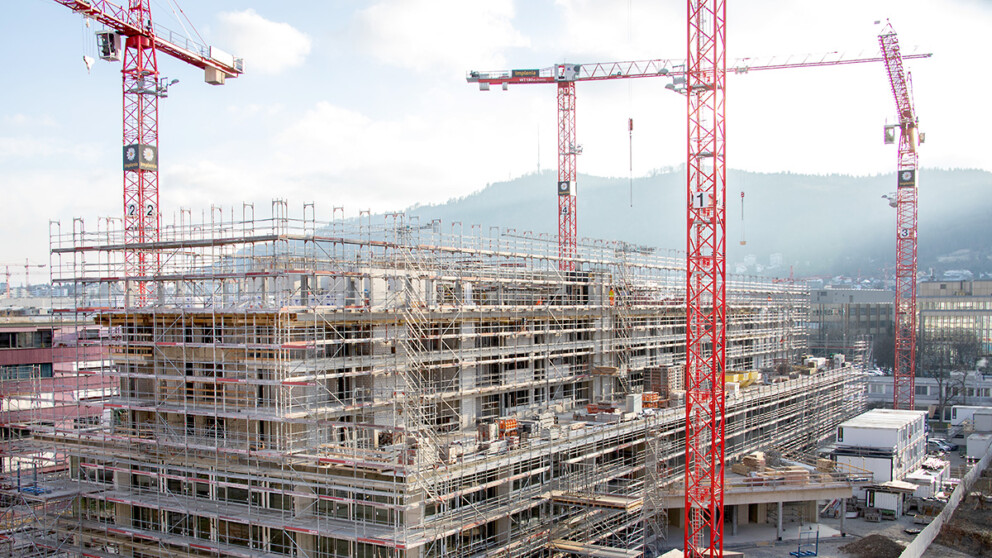Kochareal mitten in der Stadt Zürich während dem Bau (Blick Richtung Süden, im Hintergrund sieht man den Hügel des Uetlibergs): Eingerüstetes, grosses Gebäude, um das mehrere rote Baukräne stehen.