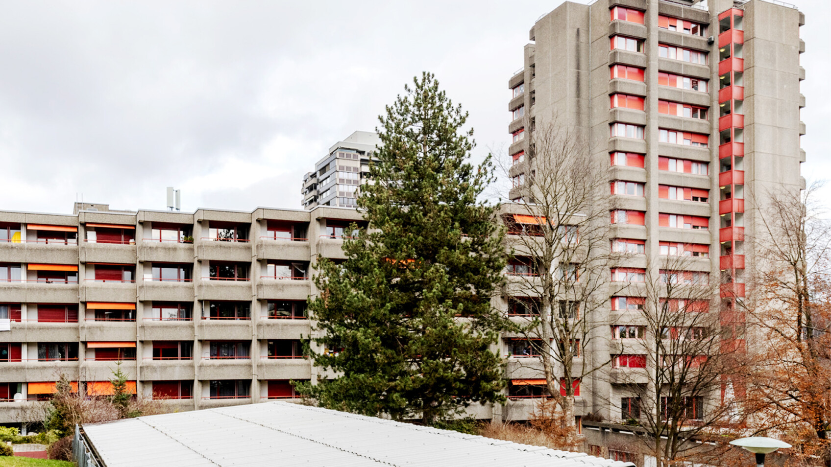 Ein großer, mehrstöckiger Wohnkomplex aus Beton mit roten Fensterakzenten und orangefarbenen Markisen, der teilweise von immergrünen und kahlen Bäumen verdeckt wird.