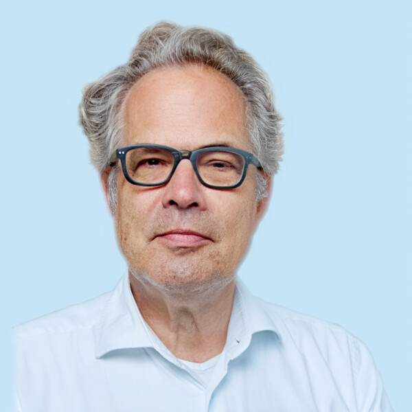 Porträt eines Mannes mit grauen Haaren, der eine Brille mit dunklem Rahmen und ein weißes Hemd trägt.