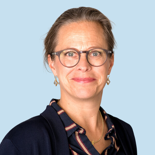 Eine Frau mittleren Alters mit Brille, die vor einem hellblauen Hintergrund lächelt.