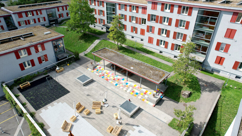 Ein Innenhof mit bunten Fliesen, Bänken, viel Grün und modernen Wohnhäusern mit roten Fensterläden, die den Bereich umgeben.