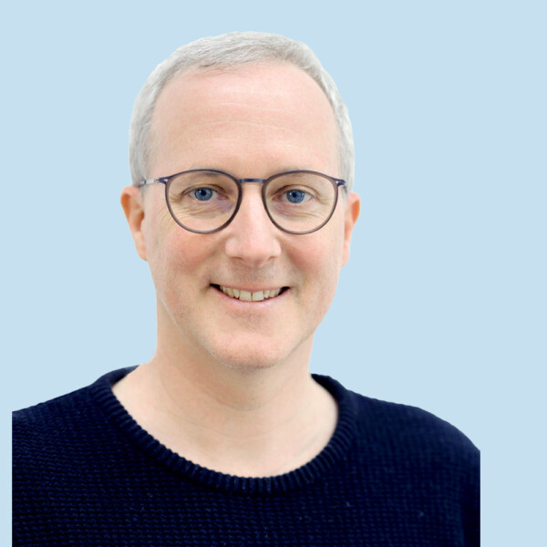Ein lächelnder Erwachsener mit kurzen grauen Haaren, Brille und einem dunklen Pullover vor einem hellblauen Hintergrund.