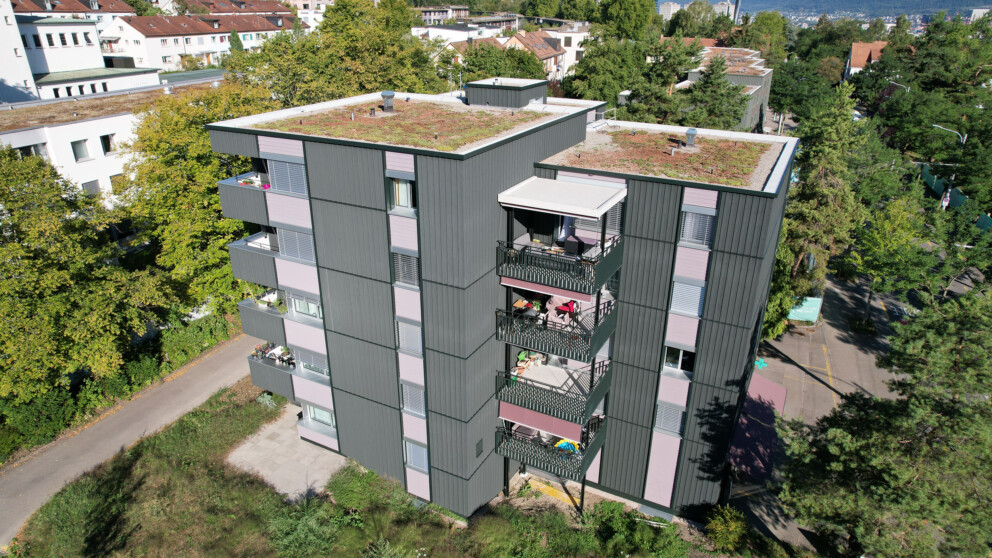 Luftaufnahme eines mehrstöckigen Wohnhauses mit dunkelgrauen Fassadenverkleidungen und begrünten Dächern, umgeben von Bäumen und Wohnhäusern.