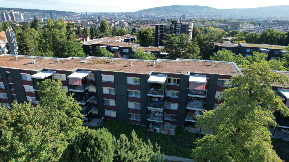 Aufnahme aus großer Höhe eines modernen Mehrfamilienhauses mit Balkonen und einem begrünten Dach, gelegen in einem Wohngebiet mit Blick auf die Stadt und die Berge in der Ferne.