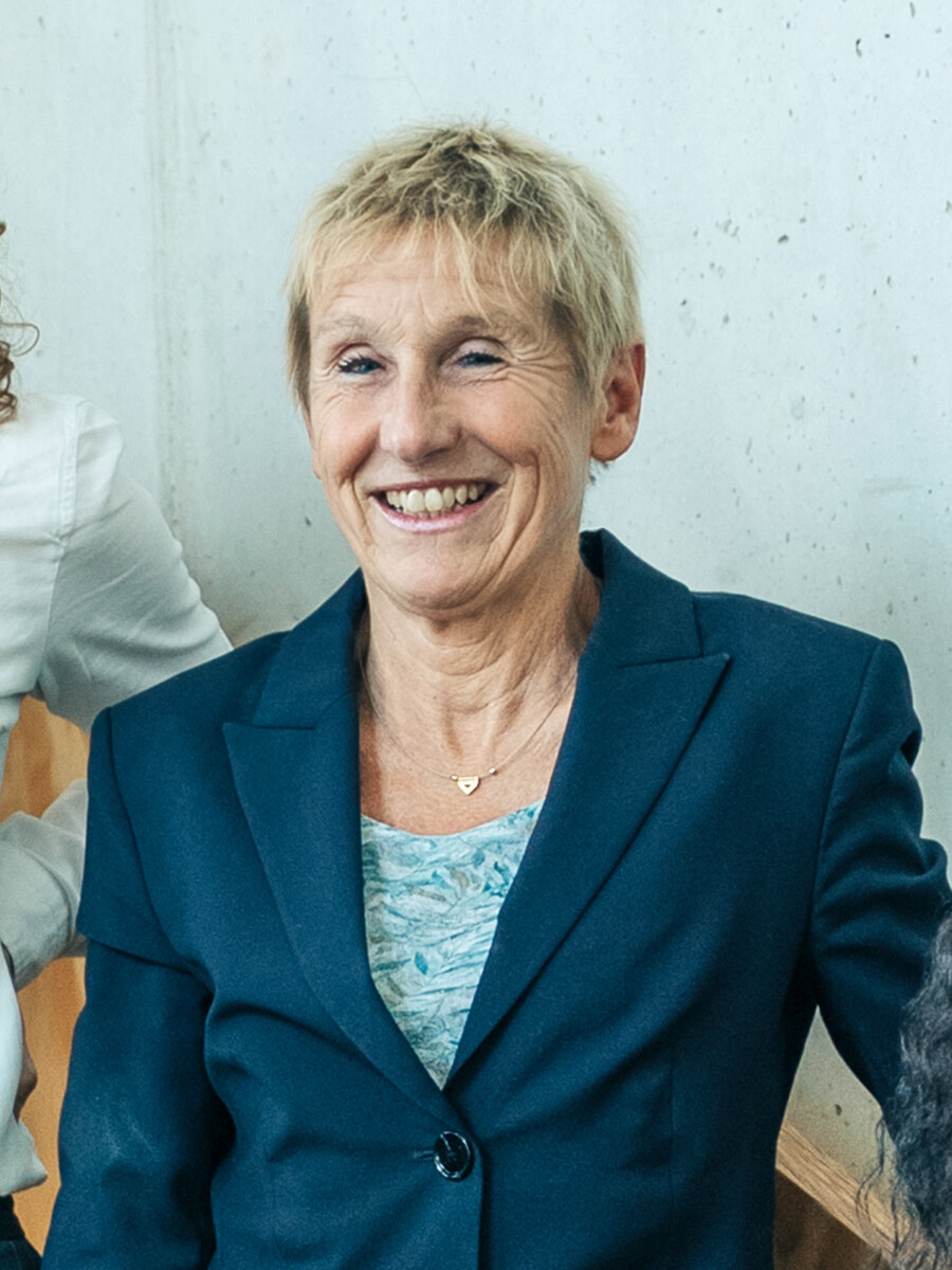 Eine ältere Frau mit kurzen blonden Haaren, die einen blauen Blazer und ein gemustertes Oberteil trägt, lächelt, während sie an einer hellen Wand sitzt.