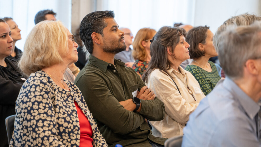 Eine bunt gemischte Gruppe von Menschen sitzt in einem Hörsaal und hört aufmerksam einer Präsentation zu. Sie sind konzentriert, manche verschränken die Arme. Der Raum ist sanft beleuchtet.