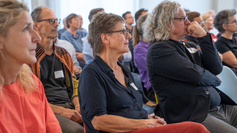 Eine Gruppe sitzender Menschen hört aufmerksam einem Vortrag zu. Die meisten tragen legere Kleidung, einige tragen Namensschilder. Im Vordergrund sitzt eine Frau in marineblauem Hemd und mit Brille neben einem Mann in schwarzer Jacke.