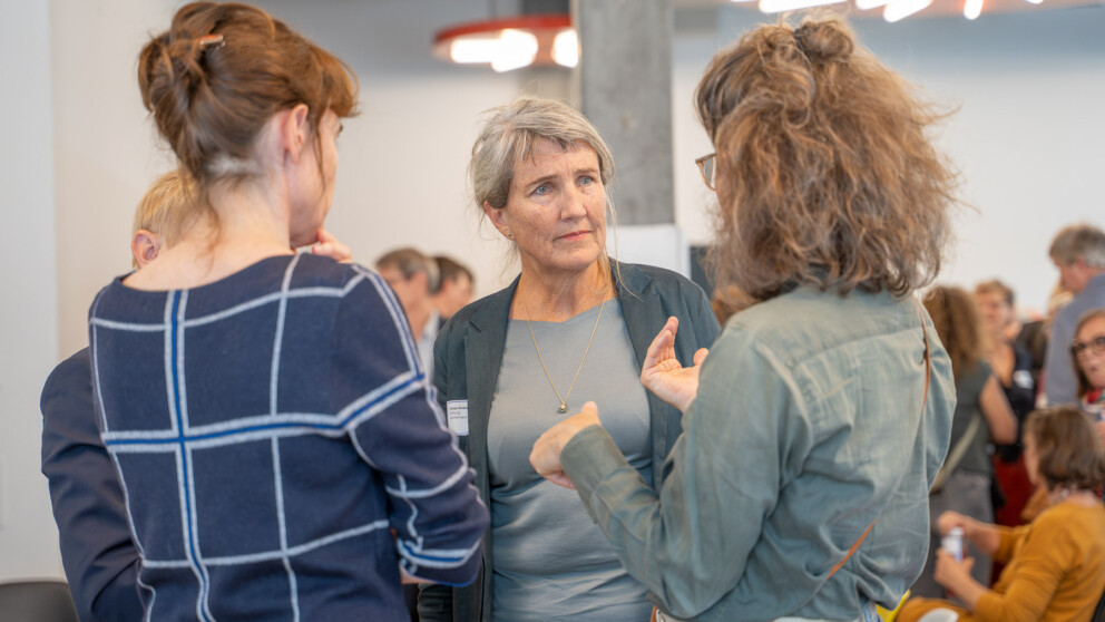 Drei Personen unterhalten sich. Der Hintergrund ist leicht verschwommen und zeigt weitere Teilnehmer und eine helle Lampe. Sie sind leger gekleidet.