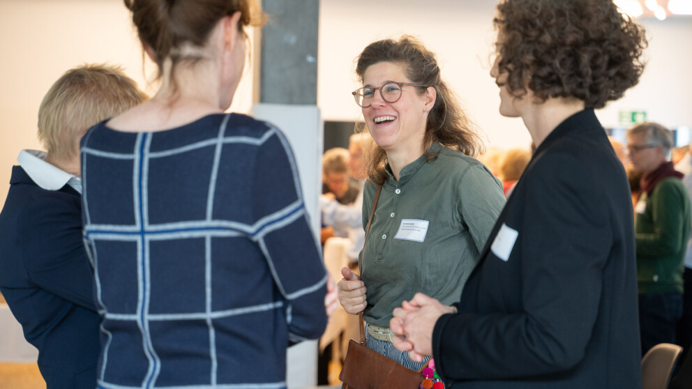 Vier Personen unterhalten. Eine Person lächelt und trägt eine Brille. Alle haben Namensschilder. Der Hintergrund ist unscharf und zeigt weitere Personen, die miteinander interagieren.
