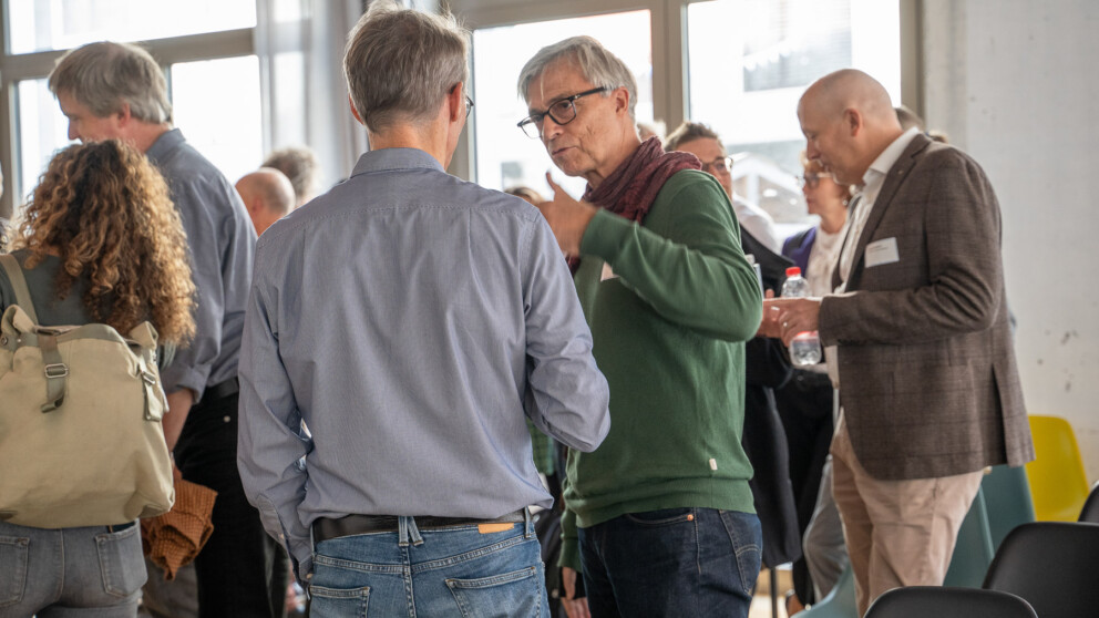 Eine Gruppe von Menschen unterhält sich in einem Raum mit großen Fenstern. Im Vordergrund sprechen zwei Männer, einer trägt einen grünen Pullover und der andere ein blaues Hemd. Im Hintergrund mischen sich andere unter die Leute.