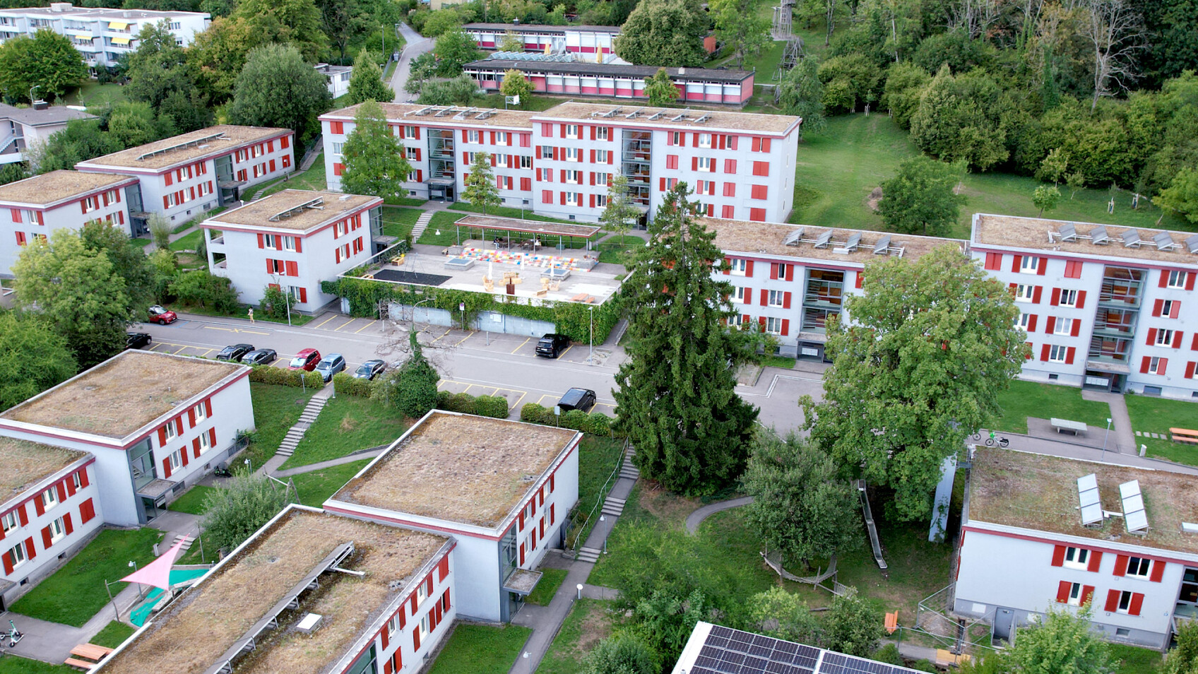 Luftaufnahme mehrerer weißer Gebäude mit roten Zierleisten, umgeben von grünen Bäumen und Rasenflächen, mit einem Swimmingpool in der Mitte.