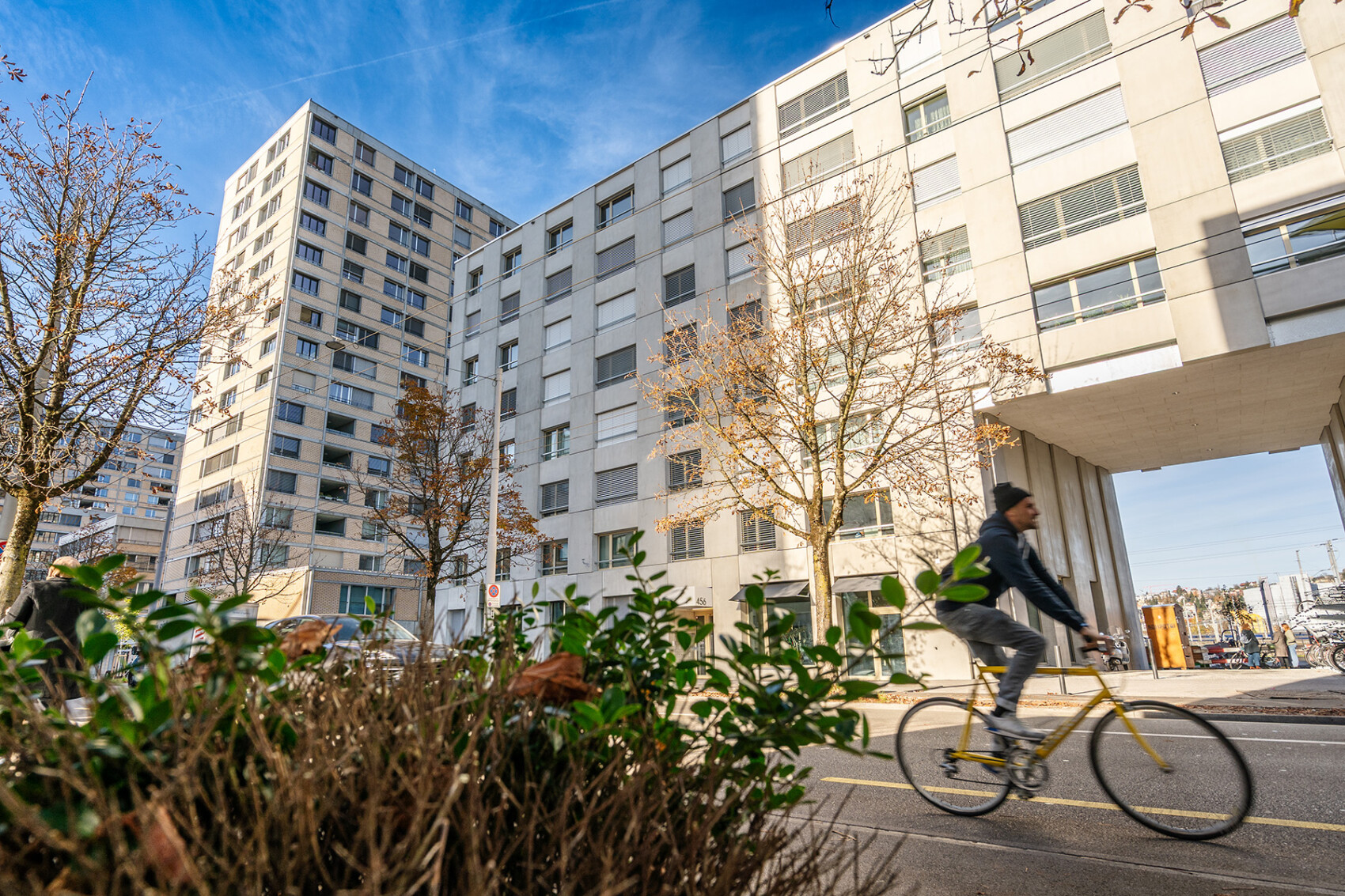 An einem sonnigen Tag mit blauem Himmel und kahlen Bäumen fährt eine Person mit dem Fahrrad an modernen, mehrstöckigen Wohnhäusern vorbei.