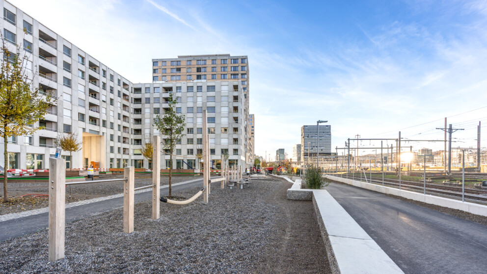 Moderne Wohngebäude neben einem minimalistischen Spielplatz mit Holzkonstruktionen, angrenzend an eine Eisenbahnstrecke unter blauem Himmel.