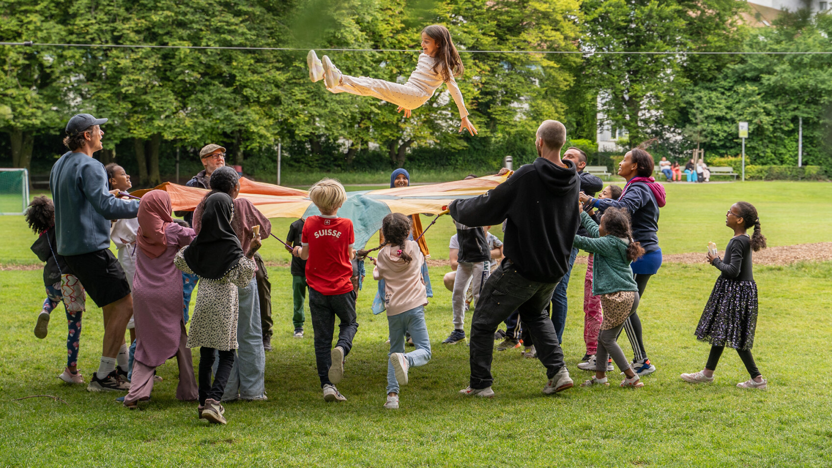 Grosse Gruppen von Kindern und Erwachsenen vergnügt sich mit einem Sprungtuch auf grüner Wiese.