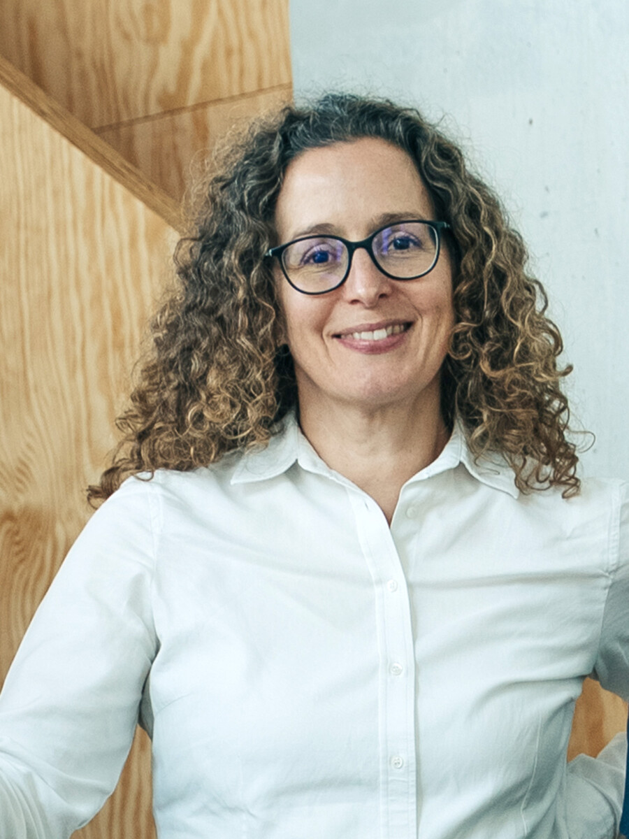 Eine Frau mit lockigem Haar und Brille, die ein weißes Hemd trägt, steht in einem Haus vor einem Hintergrund aus hellem Holz und Beton und lächelt in die Kamera.